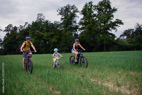 Young family with little child riding bicycles on meadow in village in summer.