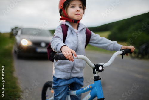 Portrait of excited little girl riding bike on road with car behind her, road safety education concept.