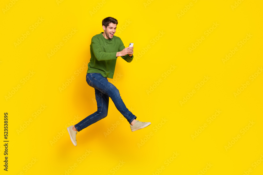 Photo of funky excited guy wear green sweater jumping writing modern device empty space isolated yellow color background