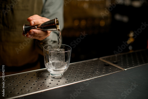 hand of bartender holds jigger and pours drink into transparent glass with ice photo