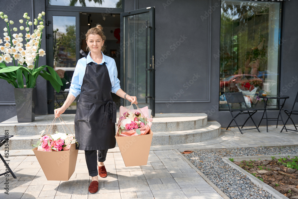 Female florist in apron carries huge package with luxurious bouquets - obrazy, fototapety, plakaty 