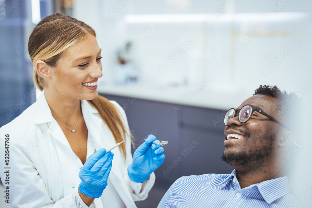 Smiling young man sitting in dentist chair while doctor examining his teeth. African American young man having a visit at the dentist's. He is sitting on chair at dentist office in dental clinic. Smiling young man sitting in dentist chair while doctor examining his teeth. African American young man having a visit at the dentist's. He is sitting on chair at dentist office in dental clinic.