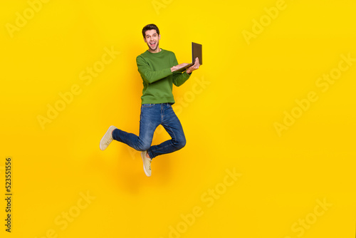 Photo of pretty funky guy wear green sweater jumping high typing modern device empty space isolated yellow color background
