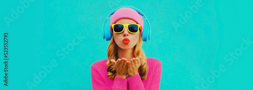 Portrait of stylish young woman in headphones listening to music blowing her lips sends sweet air kiss wearing colorful pink sweater on blue background