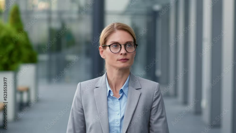 Confident middle aged business lady wearing eyeglasses walking outdoors near modern office building, follow shot Confident middle aged business lady wearing eyeglasses walking outdoors near modern office building, follow shot