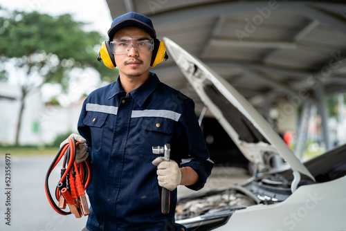 Portrait of an Asian mechanic holding a battery voltage meter in a car at a service station. Engine maintenance and repair concept. Checking the voltage level in the car battery