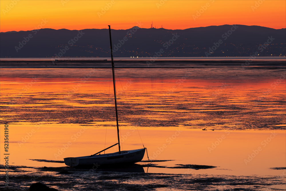Paesaggio di mare con una barca a vela ormeggiata sulla laguna del mare della città isola di Grado alla mattina all'alba. Alba colorata di arancio con i monti sullo sfondo. 