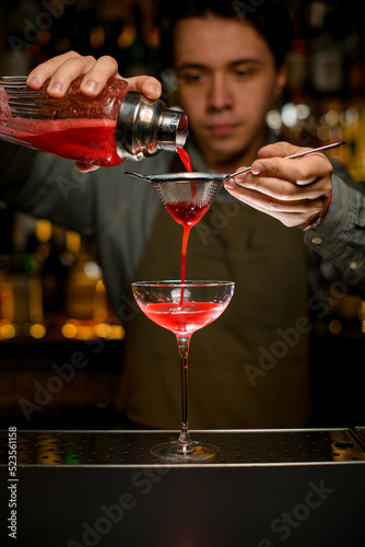 hand of male bartender holds sieve and pours cocktail through it into wine glass