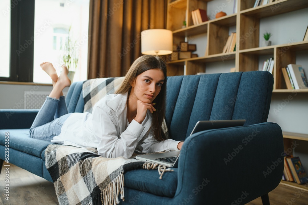 Freelancer working on laptop at home laying on comfortable beige sofa.