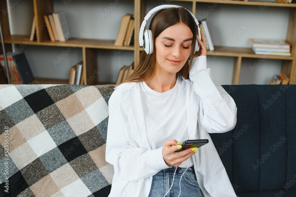 Beautiful young caucasian woman in casual clothing enjoying music and smiling while resting at home. Young woman with headphones using laptop at home Beautiful young caucasian woman in casual clothing enjoying music and smiling while resting at home. Young woman with headphones using laptop at home