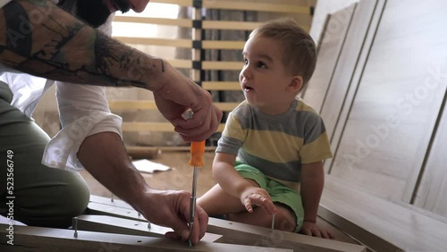 Close up view of dad and son are on the floor assembling parts for a new bed in the kids bedroom. Father screwing the screws with a screwdriver. Family activity together.