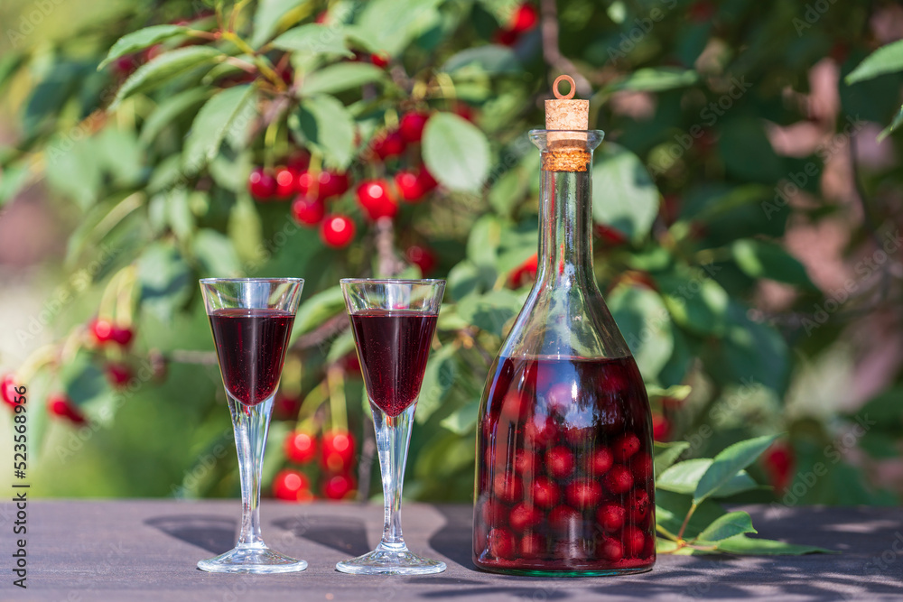 Homemade cherry brandy in two glasses and in a glass bottle on a wooden table in a summer garden