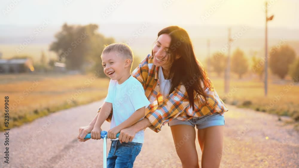 Happy face of enjoy baby boy riding scooter on asphalt road in nature outdoor. Mother runs near, holding behind wheel and keeping balance. Parent teaching son. Family active lifestyle. Toothy smile. Happy face of enjoy baby boy riding scooter on asphalt road in nature outdoor. Mother runs near, holding behind wheel and keeping balance. Parent teaching son. Family active lifestyle. Toothy smile.