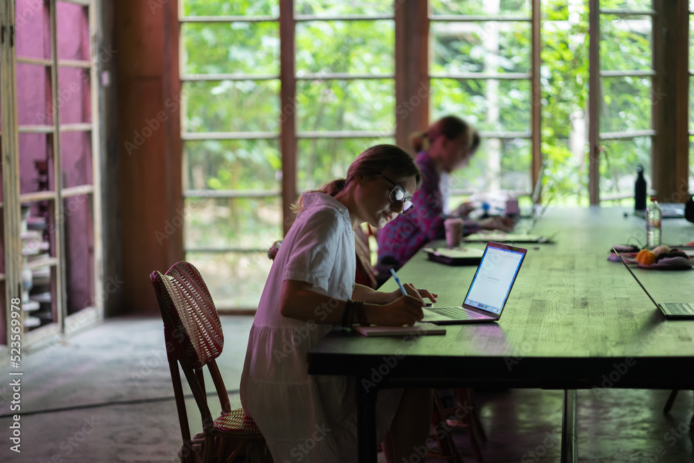 Young woman wearing glasses making notes, studying remotely in cafe, female student searching information in internet on laptop, watching webinar, learning foreign language online. Distance education Young woman wearing glasses making notes, studying remotely in cafe, female student searching information in internet on laptop, watching webinar, learning foreign language online. Distance education