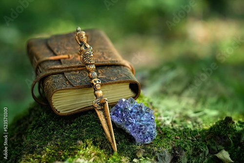 Ritual knife, amethyst druse, magic spell book on mossy stump, blurred natural green background. Phurba is three-sided knife, traditionally of Indo-Tibetan Buddhism for esoteric spiritual practice. photo