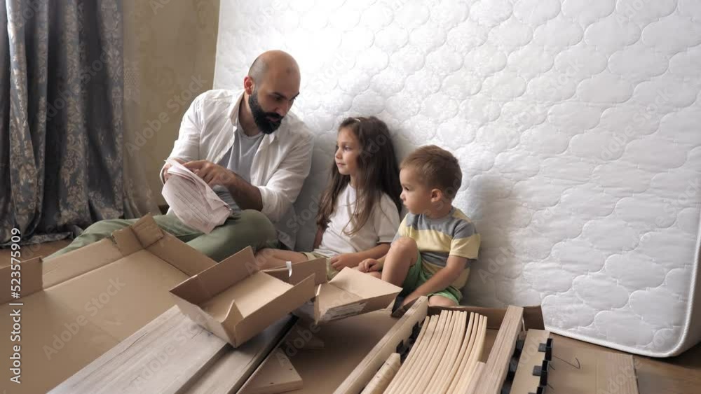 Dad with kids are on the floor reading paper instruction for a new bed in the kids bedroom. Interior structure of furniture. Helping father mounting furniture. Family activity together. Dad with kids are on the floor reading paper instruction for a new bed in the kids bedroom. Interior structure of furniture. Helping father mounting furniture. Family activity together.