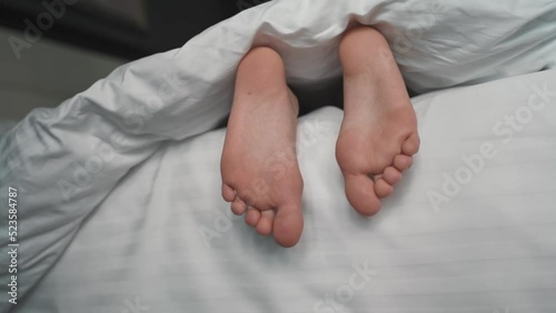 Adult child sticks legs out from under white blanket in the morning. kid of 10 years wake up in a cozy bed and waves his legs. Child legs, feet in bed. Awakening Foot Sleep Relax Concept