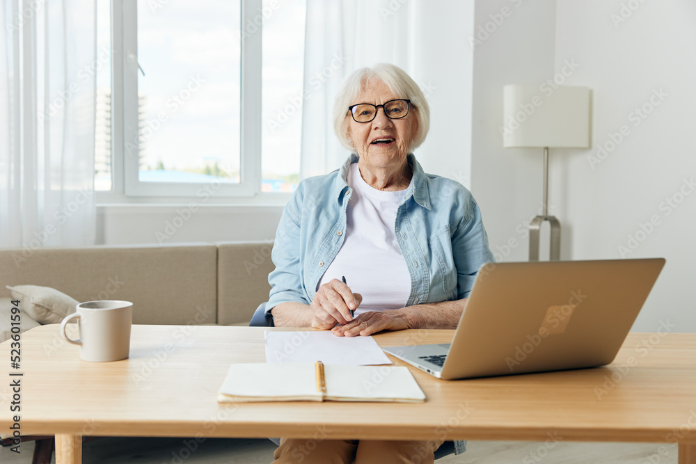 A gray-haired, beautiful, stylish, serious lady works from home sitting at her desk with a laptop and notebooks for notes, in comfort