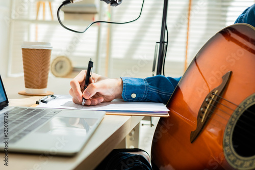 Asian young man songwriter play acoustic guitar listen song from smartphone think and write notes lyrics song in paper sit in living room at home studio. Music production at home concept.