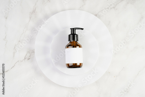 A mock-up of a brown cosmetics bottle with dispenser and white label on white round podiums on marble background, top view