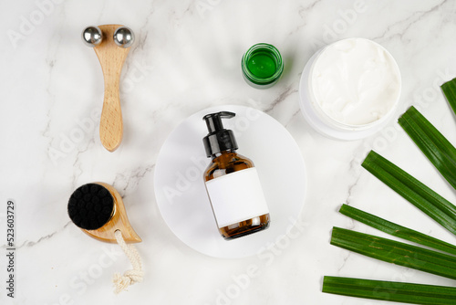 A mock-up of a brown cosmetics bottle with dispenser and white label on white round podium, round cream container, face brush, face massage roller on marble background and green palm leaf, top view