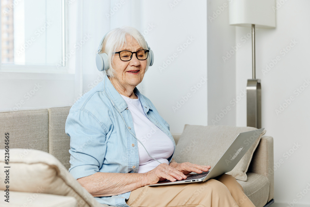 a happy, joyful old lady is sitting on a beige sofa with glasses on her face and headphones on her head working at home in a comfortable environment learning new things a happy, joyful old lady is sitting on a beige sofa with glasses on her face and headphones on her head working at home in a comfortable environment learning new things
