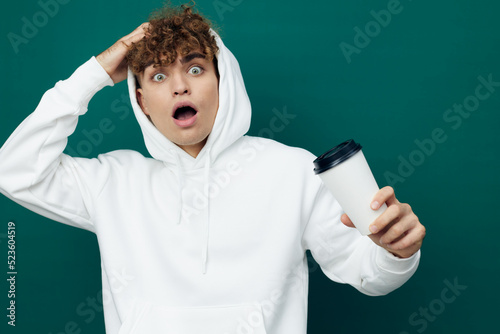 a shocked man stands on a green background in a white hoodie holding a glass in his hand and looks wide-mouthed, holding his hand on his head