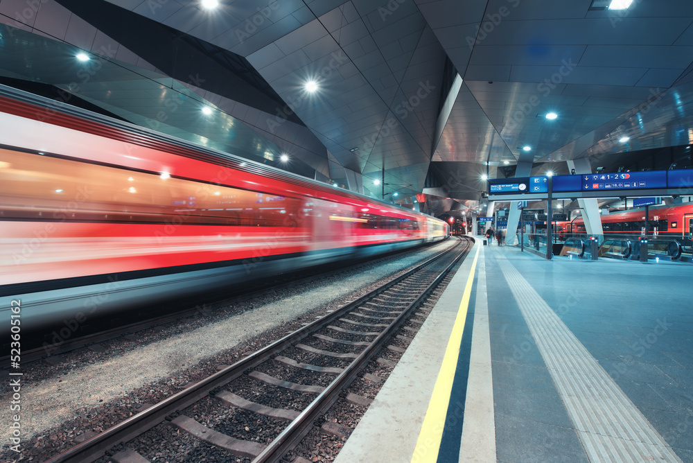 High speed train in motion on the railway station at night. Moving red modern intercity passenger train, railway platform, architecture, city lights. Modern train station in Vienna, Austria. Railroad