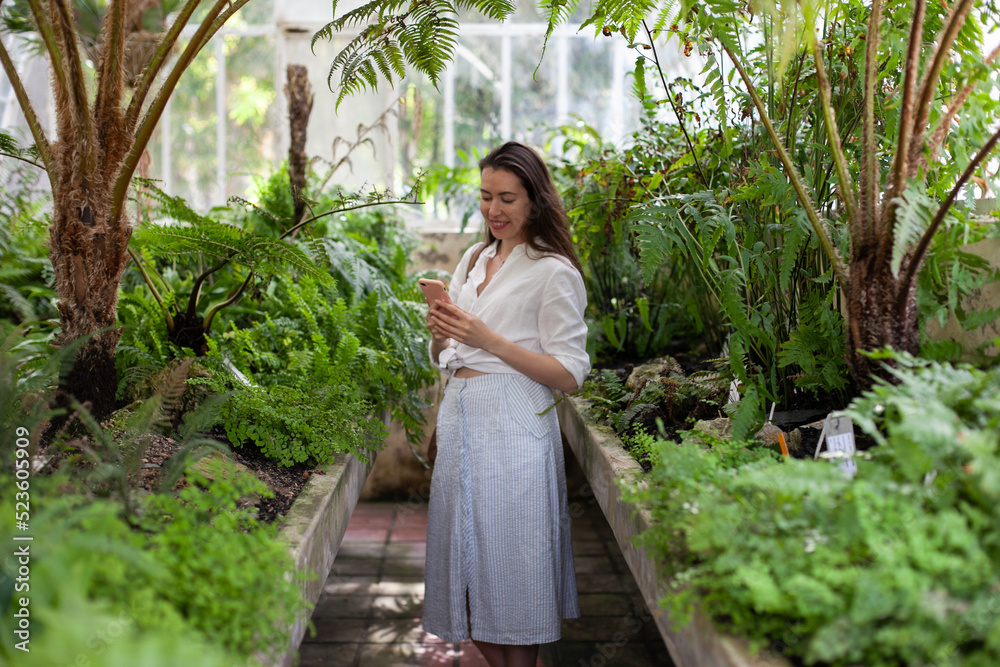 Obraz premium Young woman using mobile phone in greenhouse. Female tourist taking selfies or video chatting in nature.