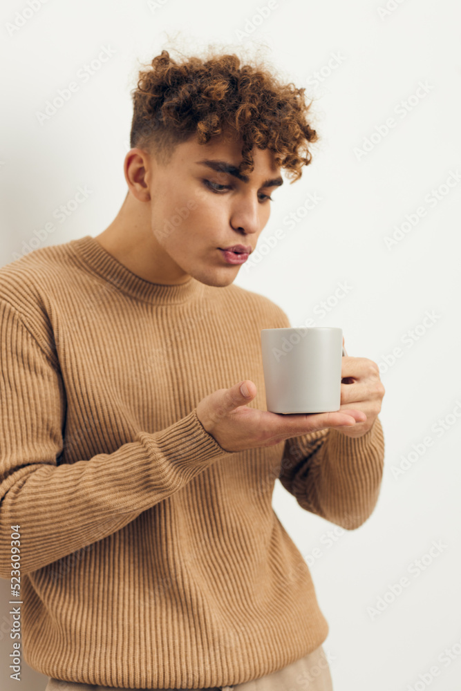 a cute, stylish, curly-haired man stands on a light background holding a white cup with a hot drink in his hand and blows on it a cute, stylish, curly-haired man stands on a light background holding a white cup with a hot drink in his hand and blows on it