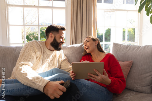 Happy caucasian couple sitting on couch in living room laughing, using smartphone and tablet