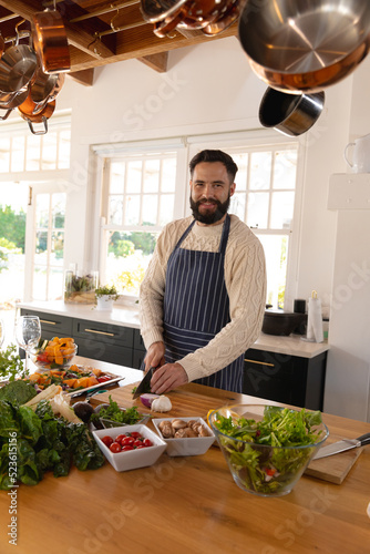 Portrait of happy caucasian man with beard chopping vegetables wearing apron in kitchen