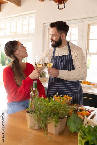 Happy caucasian couple preparing food in kitchen, talking and making a toast with glasses of wine
