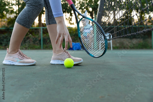 Low section of woman holding tennis racket picking up ball on outdoor tennis court, with copy space