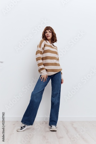 a full-length photo of a beautiful, stylish woman standing on a light background in a striped sweater and blue jeans posing in a relaxed manner