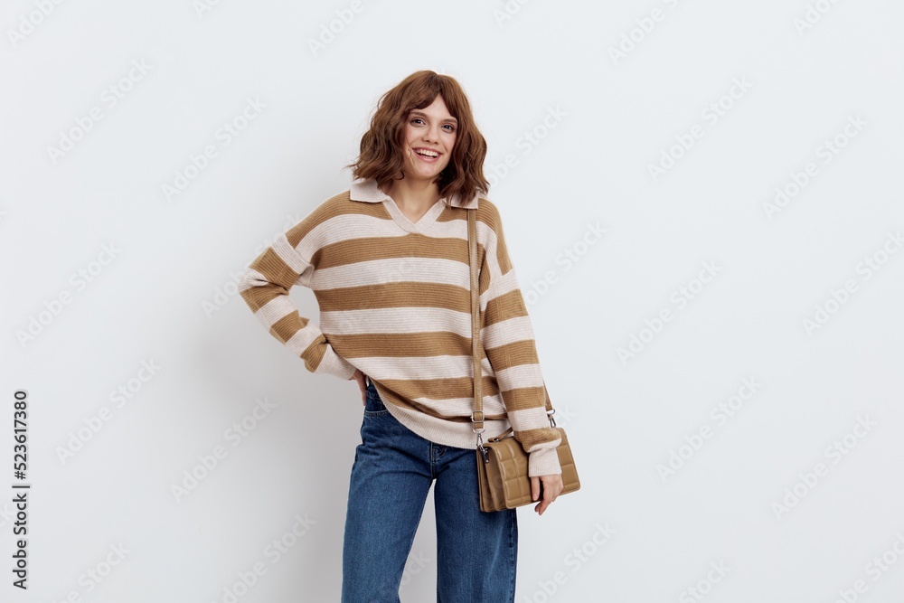 a pretty sweet woman is standing on a light background in a striped sweater and a bag on her shoulder standing half sideways with one hand on her hip lifting the sweater and lowering the other hand a pretty sweet woman is standing on a light background in a striped sweater and a bag on her shoulder standing half sideways with one hand on her hip lifting the sweater and lowering the other hand