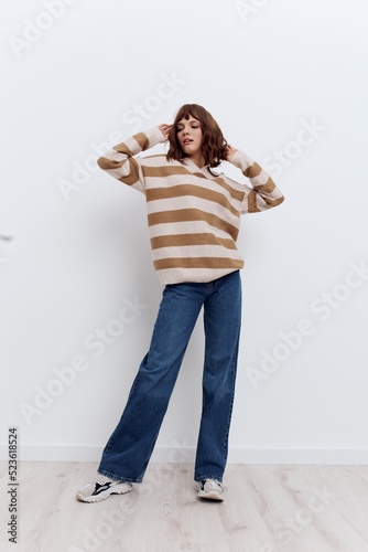 a full-length photo of a beautiful, stylish woman standing on a light background in a striped sweater and blue jeans, holding her hands on her head, spreading her elbows to the sides