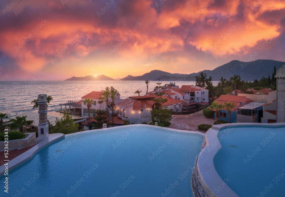 Beautiful swimming pool, sky with pink clouds at colorful sunset. Blue water, sea coast, architecture, palm trees in summer. Luxury resort. Oludeniz, Turkey. Landscape with pool, houses, orange roofs