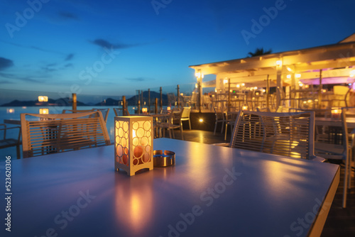 Fototapeta Naklejka Na Ścianę i Meble -  Beautiful lamp on the table in cafe on sea coast at night in summer. Landscape with candle and blurred background with chairs and tables, lights. Empty cozy cafe outdoor with illumination. Travel