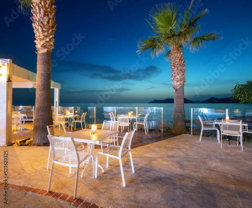 Fototapeta Naklejka Na Ścianę i Meble -  Beautiful cafe on sea coast at night in summer. Landscape with chairs and tables, lamps, lights, palm tree, water, blue sky at dusk. Empty cozy cafe outdoor with illumination in Oludeniz, Turkey