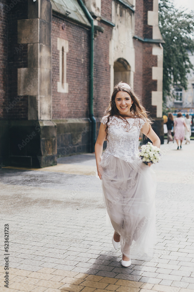 Beautiful happy bride in stylish vintage dress running in sunny street on background of church. Emotional smiling bride with wedding .bouquet of roses and lavender. Provence wedding