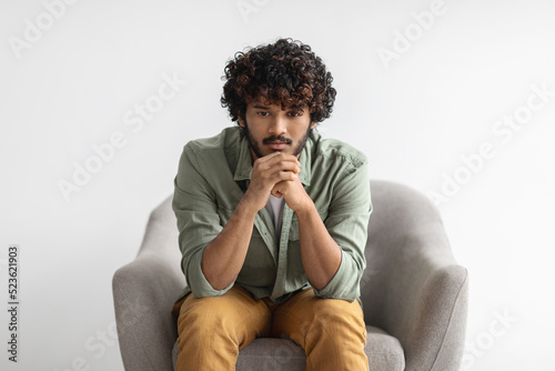 Pensive indian guy sitting in armchair over white