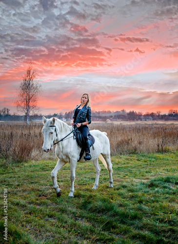 young beautiful blond smiling woman with long hair riding a white horse with blue eyes in autumn field 