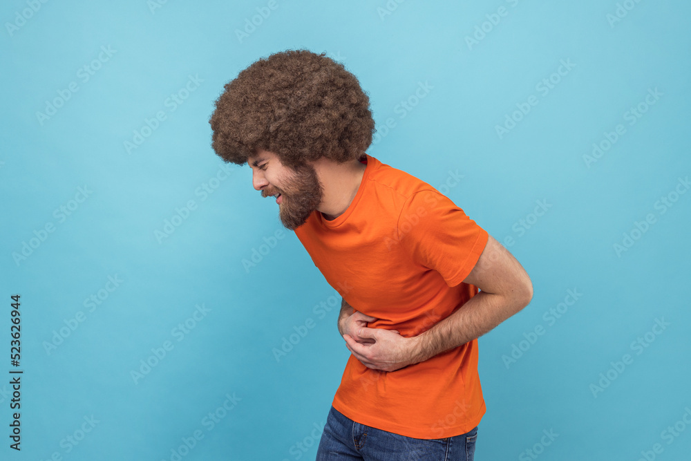 Stomach pain. Side view of sick sad unhealthy man with Afro hairstyle wearing orange T-shirt standing and holding his painful belly. Indoor studio shot isolated on blue background. Stomach pain. Side view of sick sad unhealthy man with Afro hairstyle wearing orange T-shirt standing and holding his painful belly. Indoor studio shot isolated on blue background.