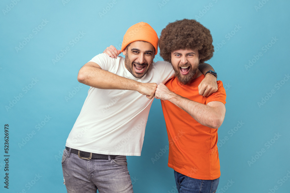 Fototapeta premium Yes we did. Portrait of two handsome happy excited young adult hipster men celebrating triumph together, showing bro fist, power five sign. Indoor studio shot isolated on blue background.