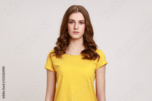 Portrait of pretty serious bossy brown haired woman of young age in yellow casual T-shirt, looking at camera with suspicion and accusing. Indoor studio shot isolated on gray background.