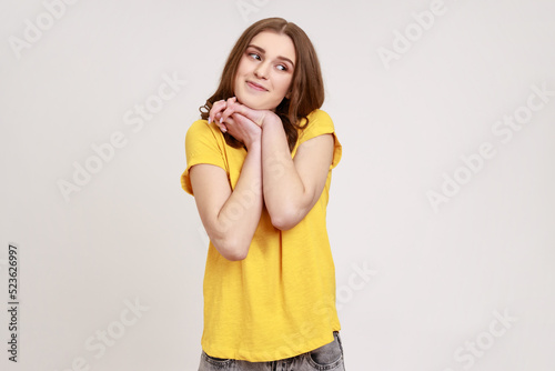 Portrait of charming inspired woman of young age with wavy hair wearing yellow T-shirt looking away with pensive expression, dreaming pleasant thoughts. Indoor studio shot isolated on gray background.