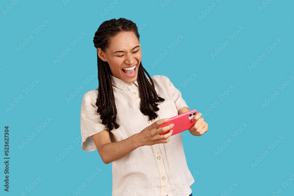 Emotional happy woman with black dreadlocks stares at smartphone camera, plays games, yelling happily while gaming smiles excited, wearing white shirt. Indoor studio shot isolated on blue background. Emotional happy woman with black dreadlocks stares at smartphone camera, plays games, yelling happily while gaming smiles excited, wearing white shirt. Indoor studio shot isolated on blue background.