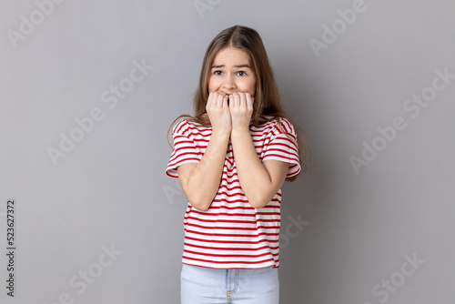 Portrait of dark haired nervous adorable little girl wearing striped T-shirt biting his fingers with shocked look, fears and phobias. Indoor studio shot isolated on gray background. photo