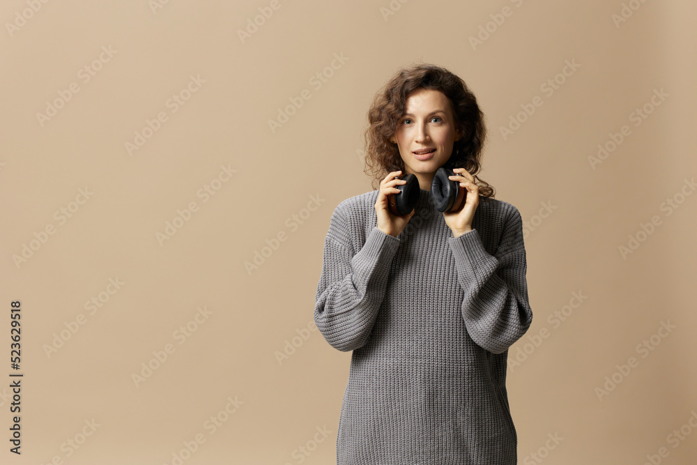Obraz premium Cheerful enjoyed pretty curly beautiful female in gray casual sweater wear headphones looks at camera posing isolated on over beige pastel background. Sound streaming platform concept. Copy space